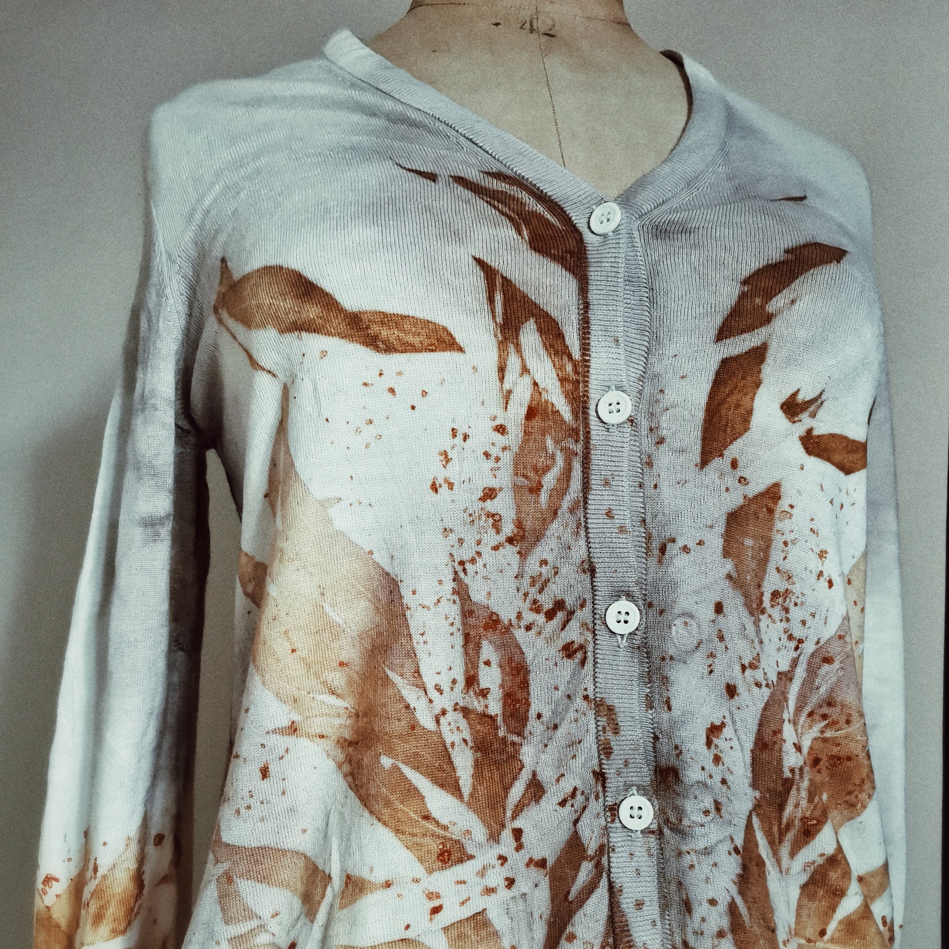 Vue de face d'un cardigan blanc boutonné sur un buste de couture. Le vêtement est orné d'une composition artistique de feuilles imprimées par contact (écoprint) aux couleurs automnales, créant un design asymétrique et organique.