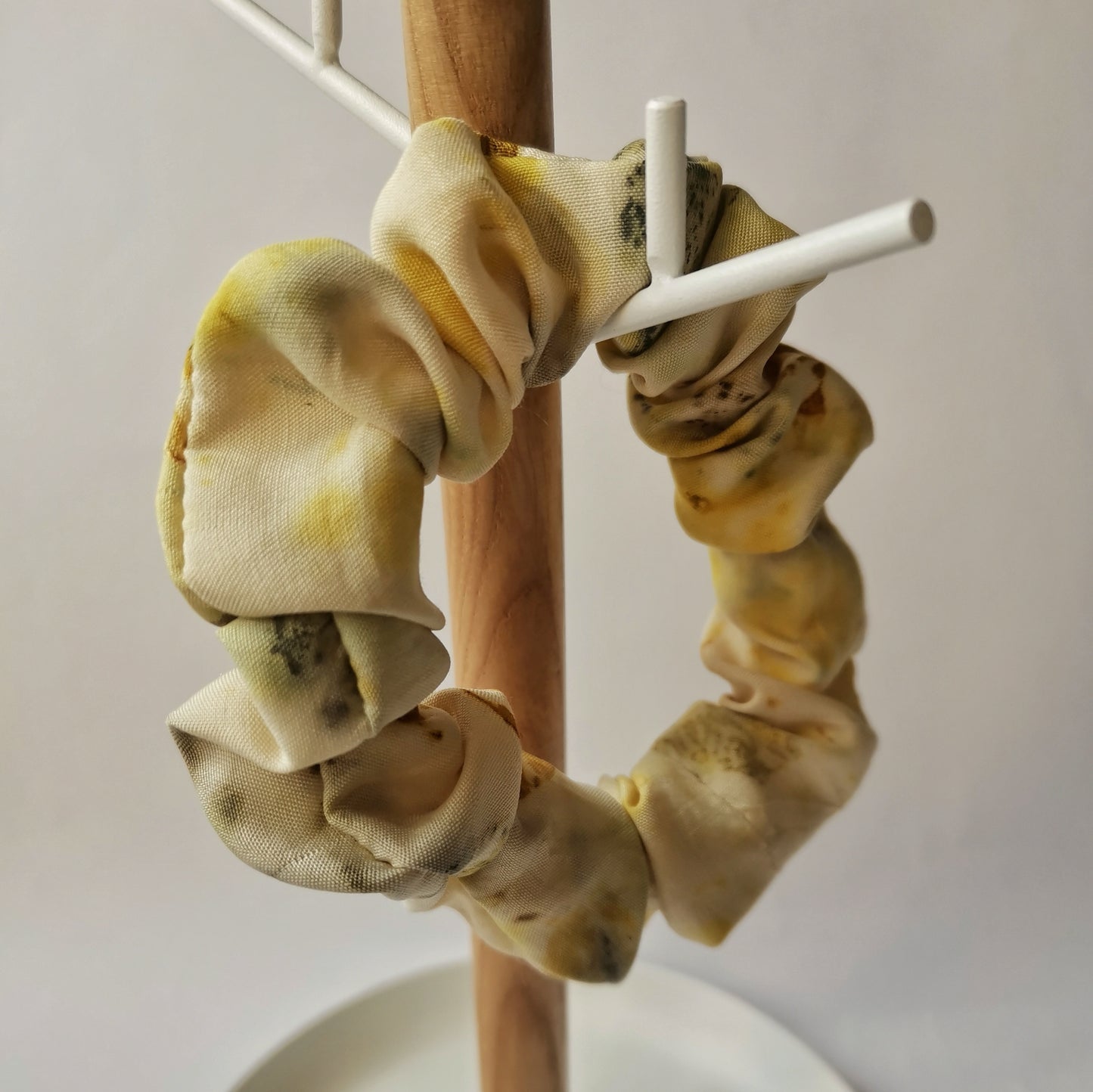 Gros plan d'un chouchou en soie jaune pâle avec des motifs botaniques délicats, présenté sur un support en bois naturel et métal blanc.