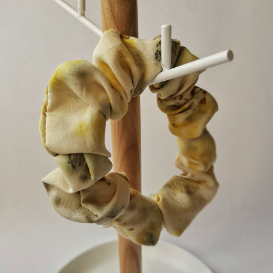 Gros plan d'un chouchou en soie jaune pâle avec des motifs botaniques délicats, présenté sur un support en bois naturel et métal blanc.