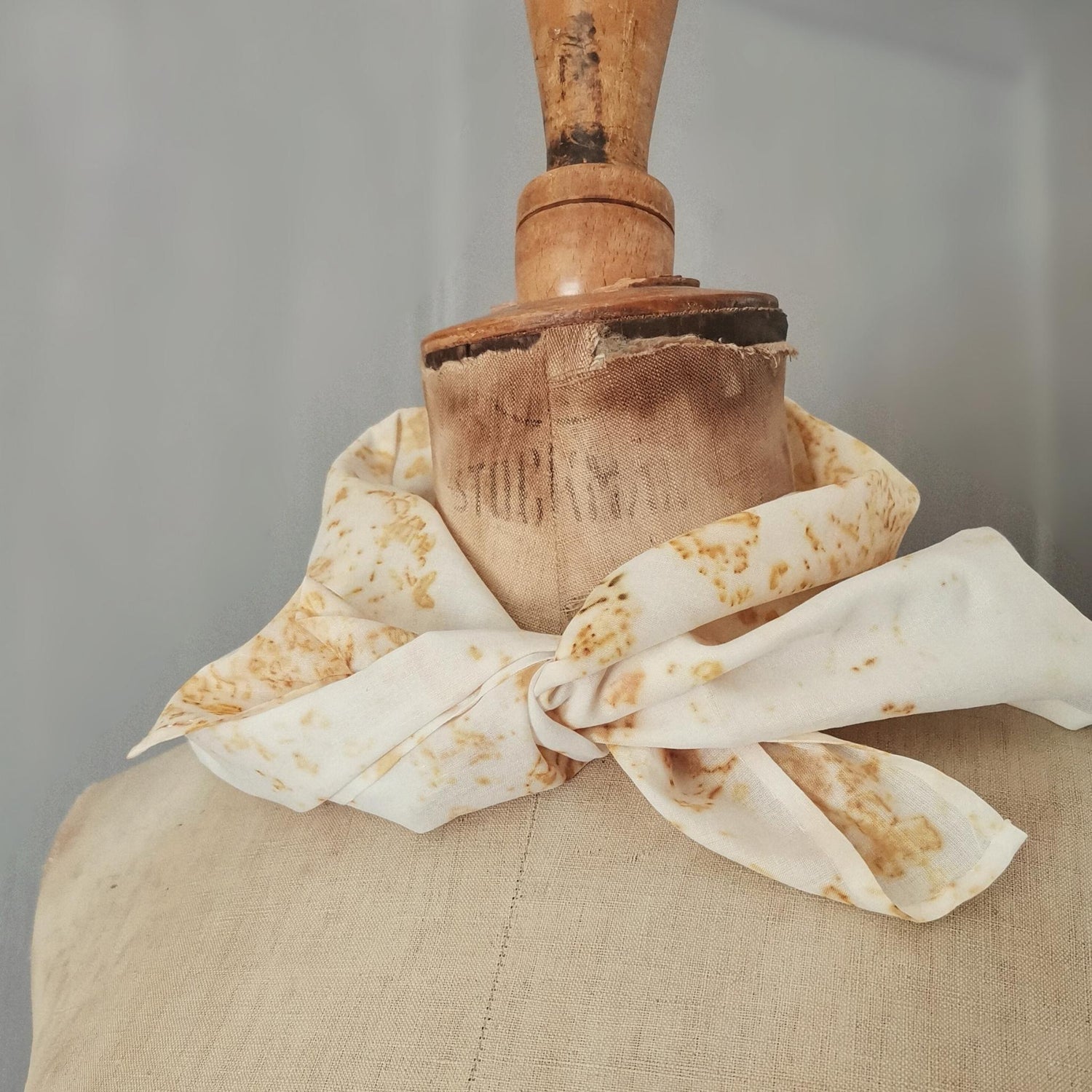 Foulard noué sur un buste de couture, confectionné en textile ancien avec des motifs botaniques jaunes obtenus par empreinte végétale. Fond gris à lattes verticales.