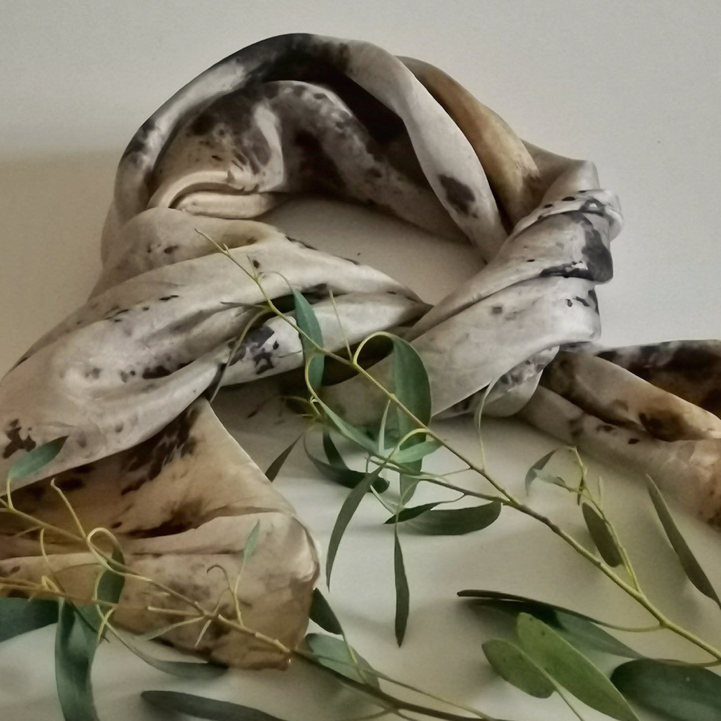 Foulard en soie upcyclée teinté artisanalement aux pigments végétaux, motifs botaniques gris et ocre, posé avec une branche d'eucalyptus sur fond clair.