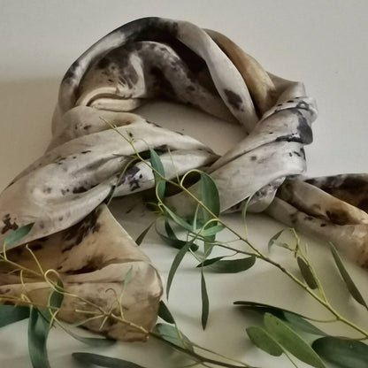 Foulard en soie upcyclée teinté artisanalement aux pigments végétaux, motifs botaniques gris et ocre, posé avec une branche d'eucalyptus sur fond clair.