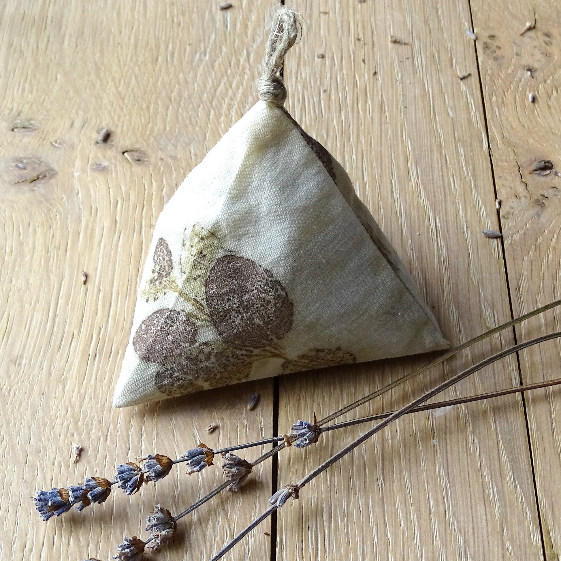Zoom sur un sachet de lavande berlingot avec une empreinte précise de feuilles d'eucalyptus brune sur coton blanc, posé sur un bois clair avec des brins de lavande séchée.