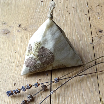 Zoom sur un sachet de lavande berlingot avec une empreinte précise de feuilles d'eucalyptus brune sur coton blanc, posé sur un bois clair avec des brins de lavande séchée.