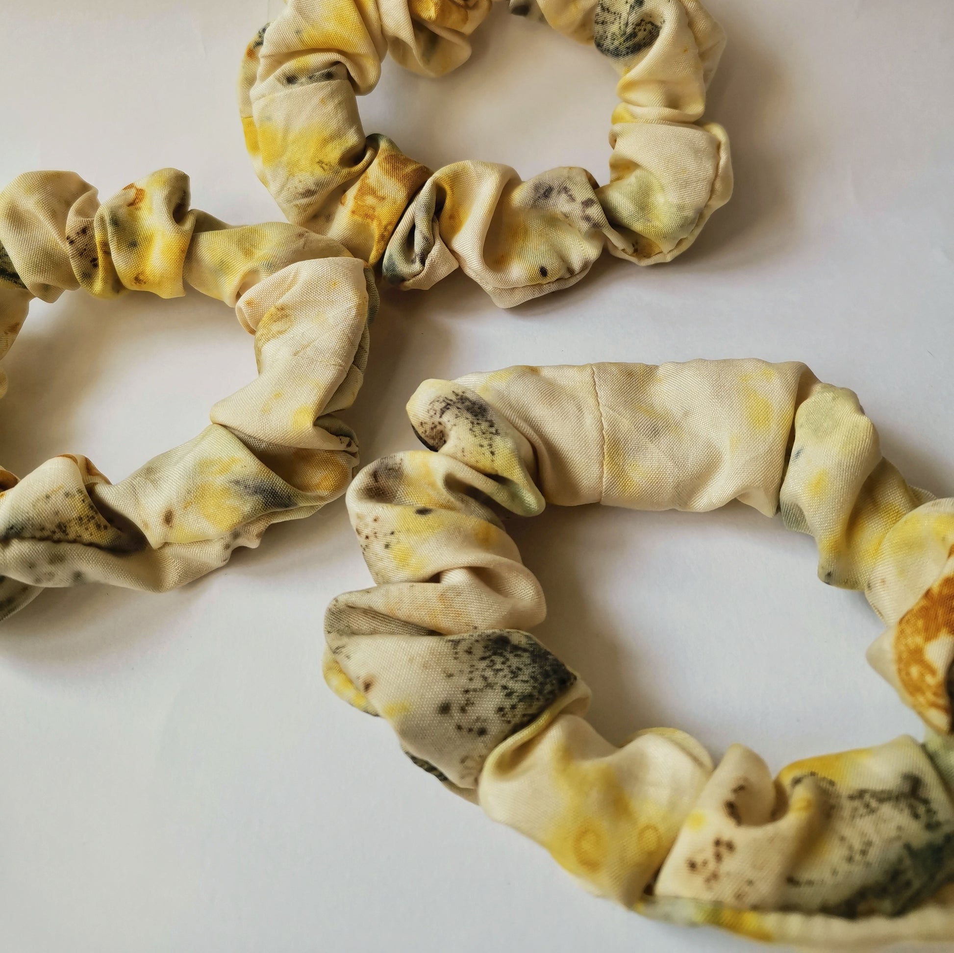 Vue rapprochée de trois chouchous faits main en tissu naturel jaune clair avec des empreintes végétales foncées, mettant en avant la texture du textile et les détails de l'impression artisanale.