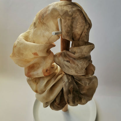 Chouchou en soie teinté naturellement suspendu à un support en bois, mettant en avant les plis et les reflets du tissu.